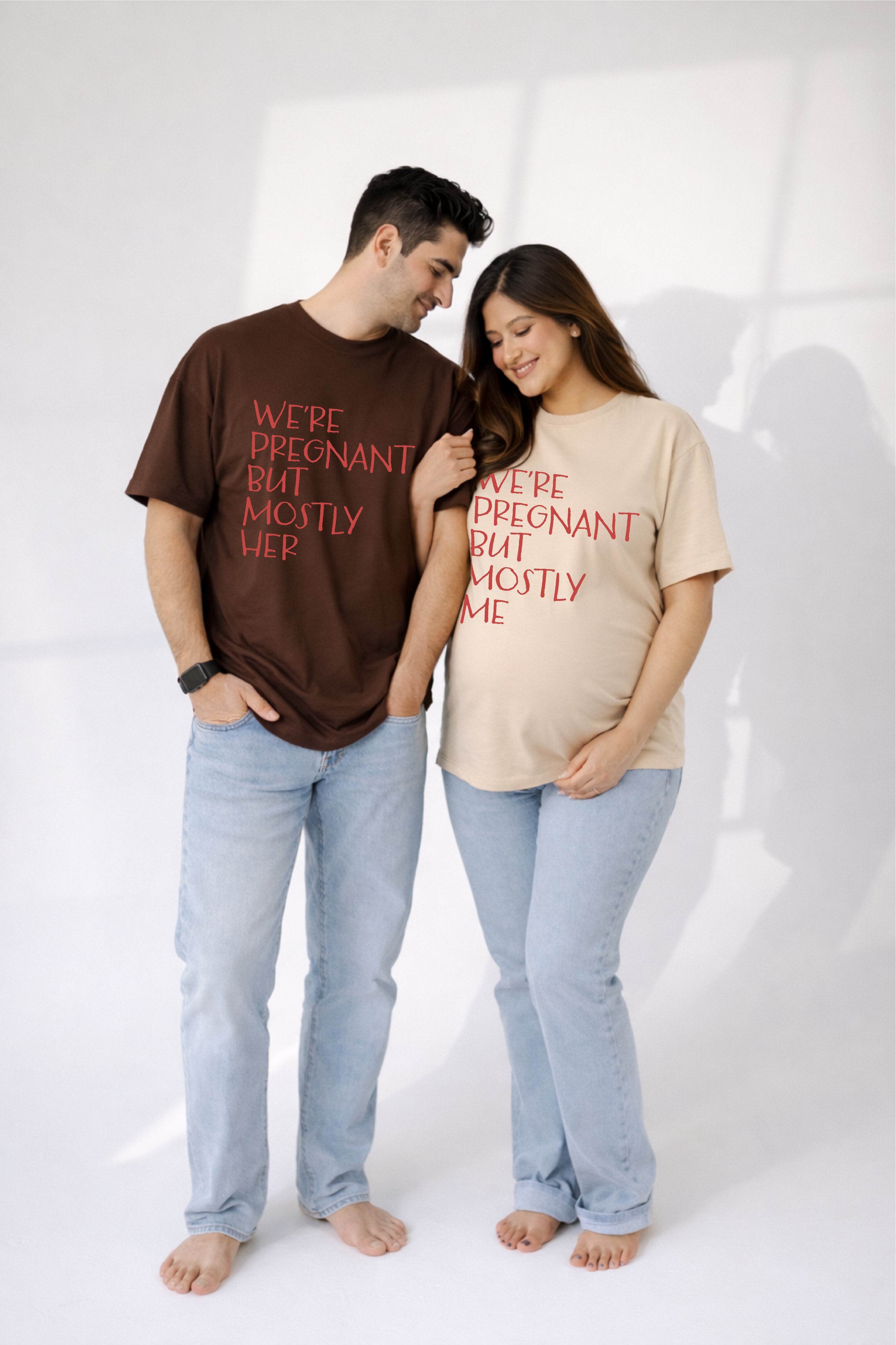 Man and woman wearing matching t-shirts with humorous pregnancy text on a white background